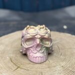 Ornate baroque-style resin skull planter with intricate scrollwork, finished in a chameleon colour-shifting shimmer of dusky pink, olive gold, emerald green, and dark gunmetal, displayed on a natural wood slice in warm sunlight.