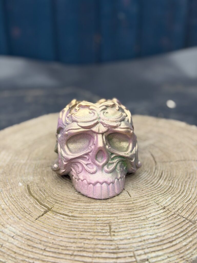 Ornate baroque-style resin skull planter with intricate scrollwork, finished in a chameleon colour-shifting shimmer of dusky pink, olive gold, emerald green, and dark gunmetal, displayed on a natural wood slice in warm sunlight.