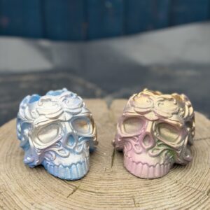Pair of ornate baroque-style resin skull planters displayed side by side on a natural wood slice — Mystic Frost in icy blue and pearl silver on the left, Enchanted Ember in dusky pink, olive gold, and emerald on the right, against a teal wooden background.
