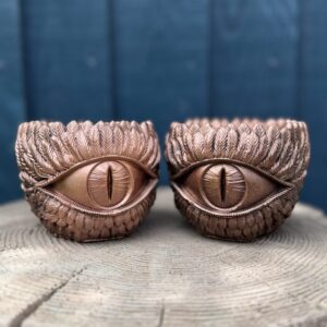 Two copper dragon eye planters side by side on a wooden log, each with a slit pupil and feathered brow, against a blue fence.