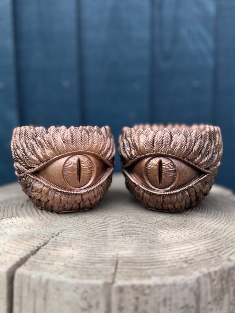 Two copper dragon eye planters side by side on a wooden log, each with a slit pupil and feathered brow, against a blue fence.