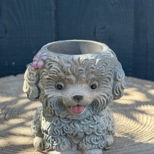 Hand-cast curly puppy planter in soft stone grey with a pink flower behind one ear and its tongue out, on a wooden log