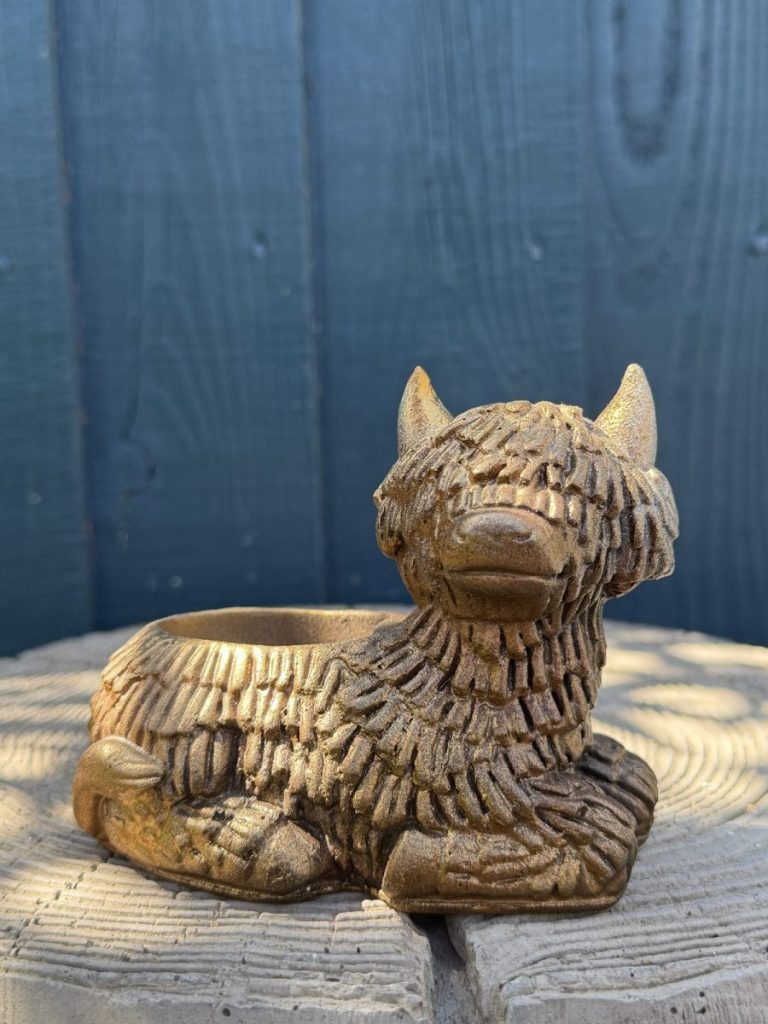 Hand-cast Highland cow planter in antique gold, shaggy coated and lying down with a planting well in its back, on a wooden log
