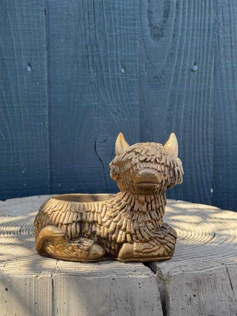 Hand-cast Highland cow planter in antique gold, shaggy coated and lying down with a planting well in its back, on a wooden log