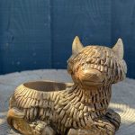 Hand-cast Highland cow planter in antique gold, shaggy coated and lying down with a planting well in its back, on a wooden log