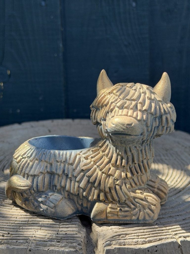 Hand-cast Highland cow planter in antique silver, shaggy coated and lying down with a planting well in its back, on a wooden log