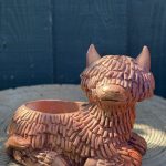Hand-cast Highland cow planter in copper rose, shaggy coated and lying down with a planting well in its back, on a wooden log