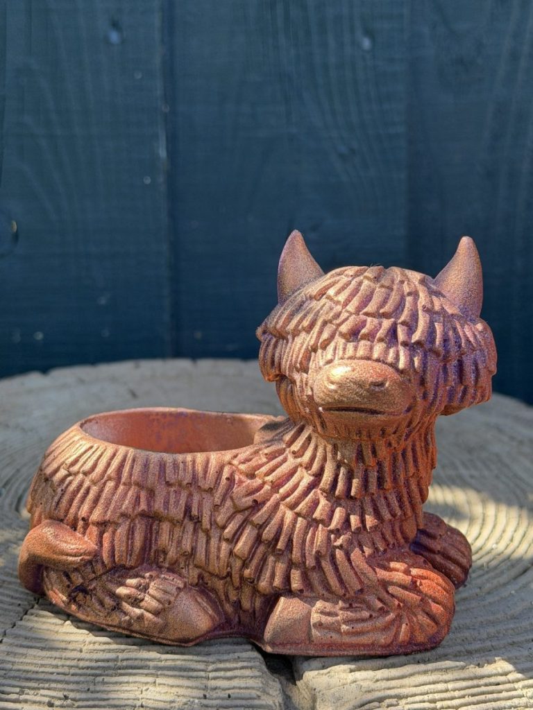 Hand-cast Highland cow planter in copper rose, shaggy coated and lying down with a planting well in its back, on a wooden log