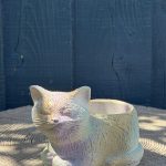 Hand-cast sleeping cat planter with a green-tinted iridescent finish, curled up with eyes closed, on a wooden log
