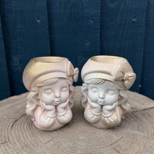 Hand-cast vintage doll planter in blush pink, wearing a beret with a bow and resting her chin on mittened hands, on a wooden log
