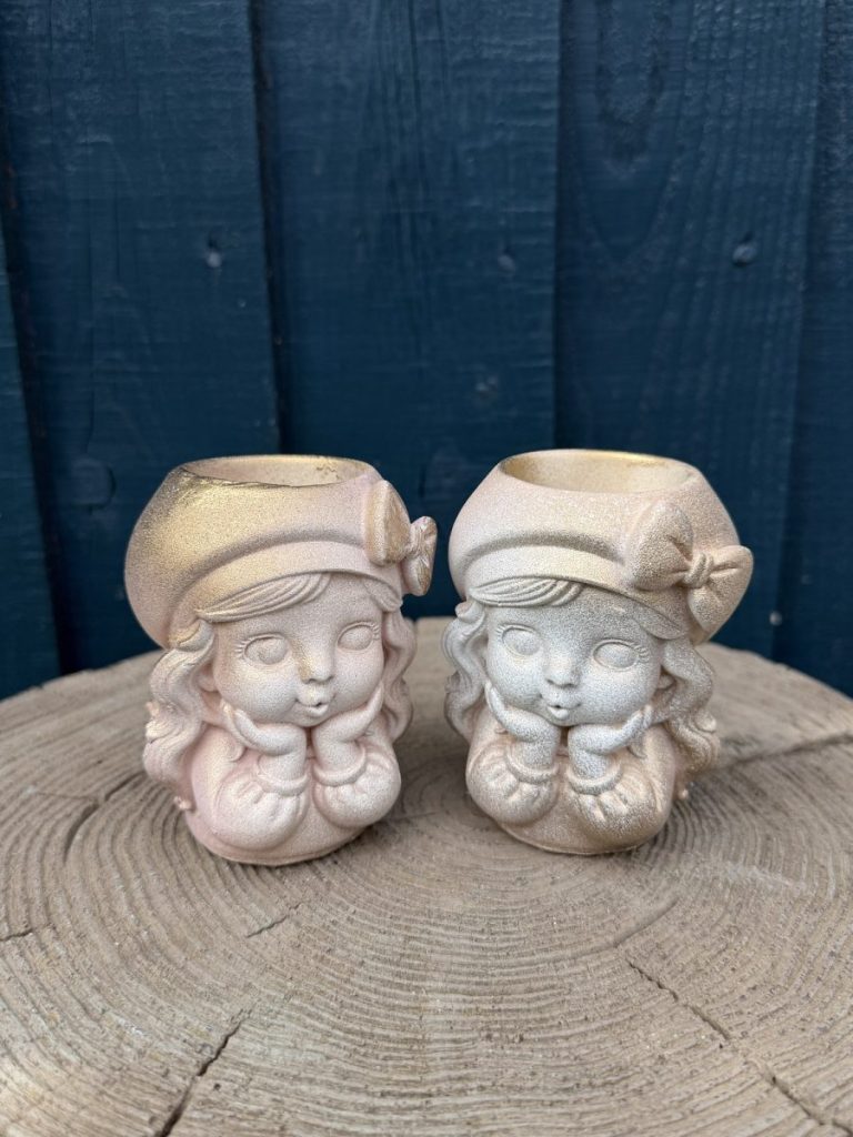 Hand-cast vintage doll planter in blush pink, wearing a beret with a bow and resting her chin on mittened hands, on a wooden log