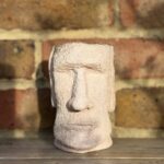 Handmade peach shimmer Moai head planter with textured cement surface glowing in sunlight on a wooden shelf in front of a red and yellow brick wall.