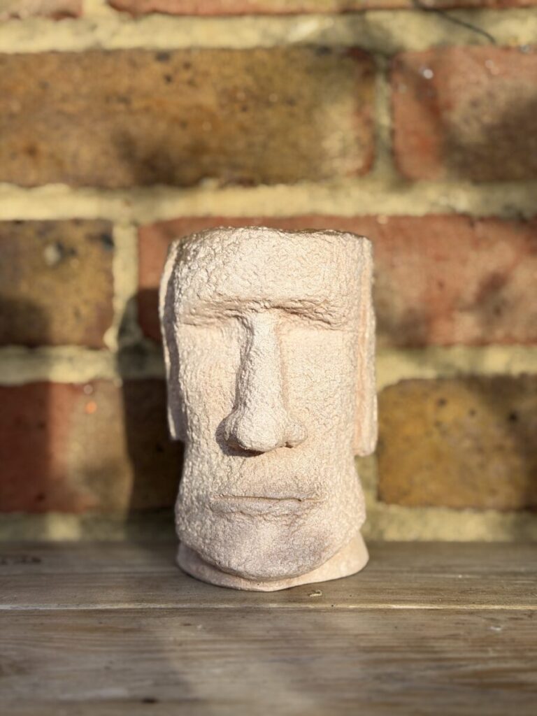 Handmade peach shimmer Moai head planter with textured cement surface glowing in sunlight on a wooden shelf in front of a red and yellow brick wall.